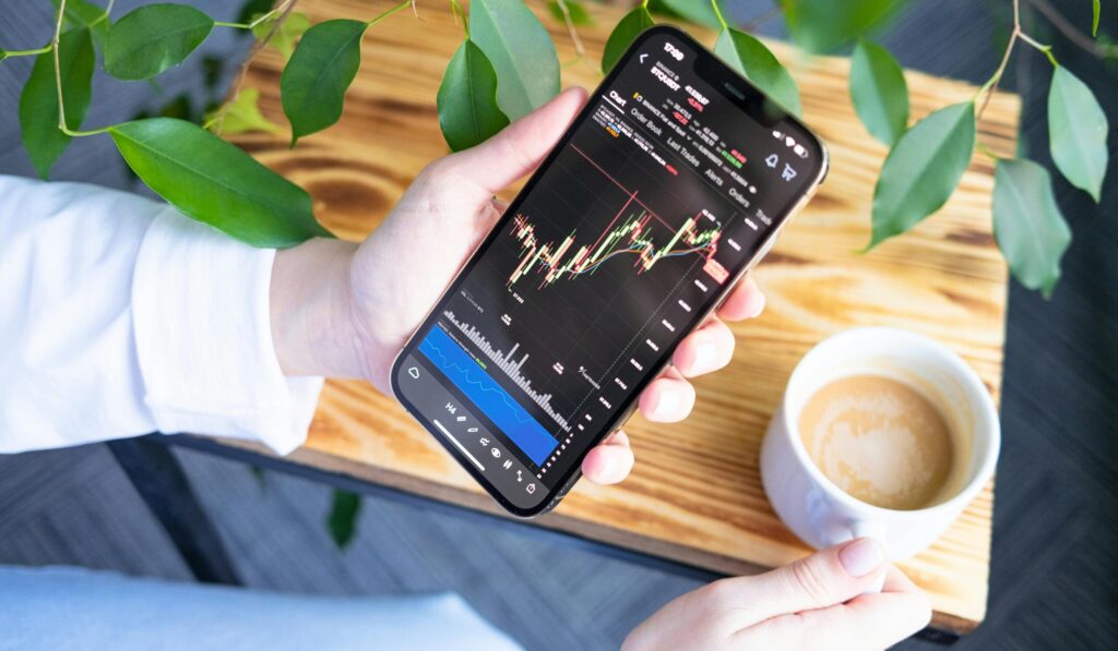 Woman holding smartphone showing stock market graph while enjoying a cup of coffee.